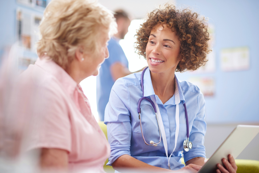 A female consultant with curly hair sits next to an elderly patient on a bench. They are talking and smiling.
