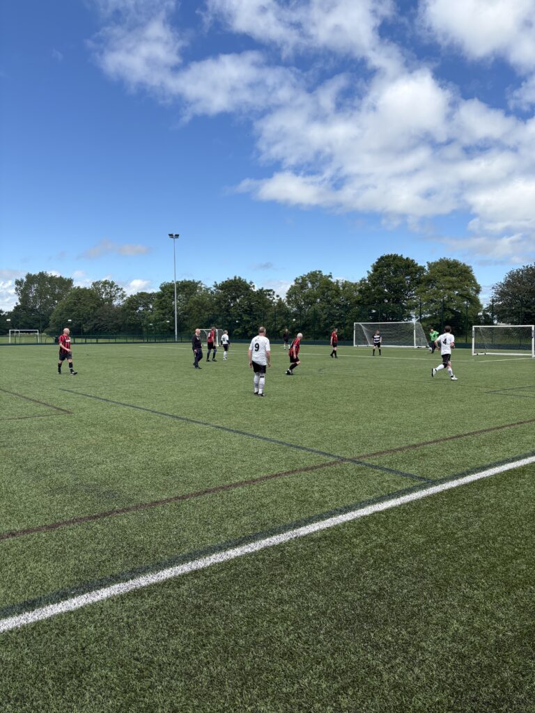 Two football teams playing football on the green pitch. One team playing in white shirts and black shorts. The other in black and red stripes with black shorts. Green trees in the background and a big blue sky with white fluffy clouds. 