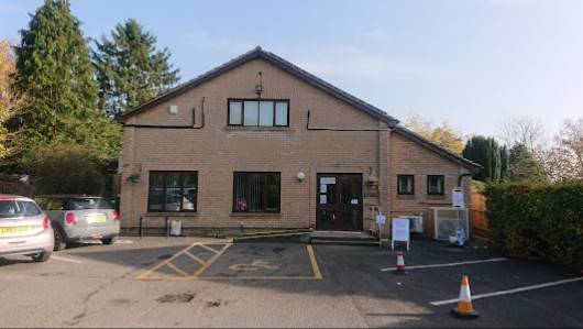A wide brown brick building with dark brown roof, window frames and doors. over looking a carpark. In front of the doors is a yellow marked disabled parking bay.