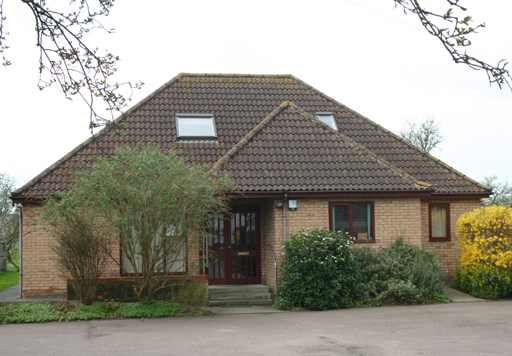 A brown brick building with dark brown roof tiles. Brown framed windows and doors. Outside is trees and green foliage surrounding the building.