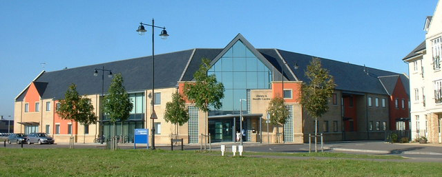 A large wide building with 5 trees Infront. The building is cream and orange brick with grey roof and glass front.