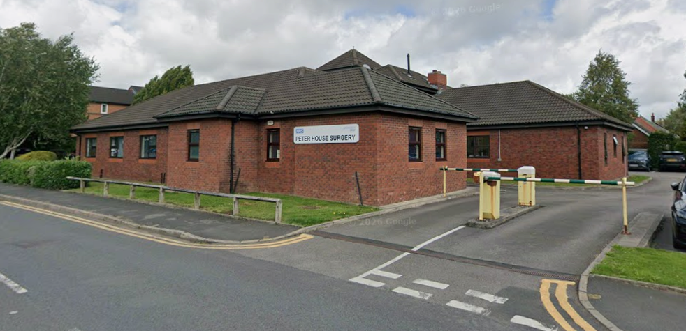 Large brown brick building with pointy grey tiled roof. White sign sharing the name Peter House Surgery