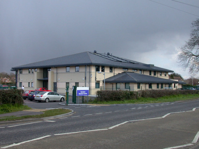 A large wide, white building with dark grey roof. The building is surrounded by a green metal fence and hedge row. At the entrance of the fence you can see a purple sign welcoming you to the centre's carpark.
