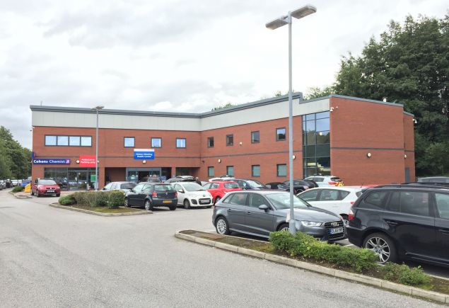 Large brown brick building with a carpark in front. A blue and white NHS sign about grey glass doors and a small chemist attached.