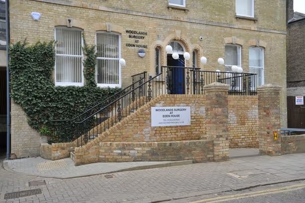 Light brick building with IVY on the front left. There is a blue door with steps leading to it from the left. A white sign on the brick shares the details of the surgery name.