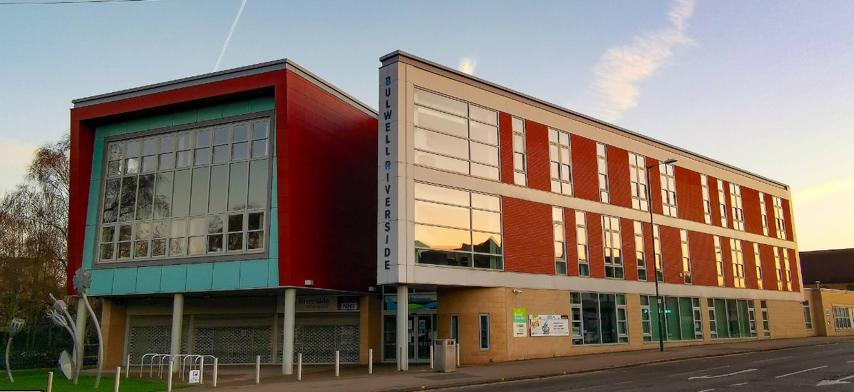 This is a large building on one side lots of glass windows inside a redbrick box and green front. On the side is a thin side of the building orange brink and windows. 
