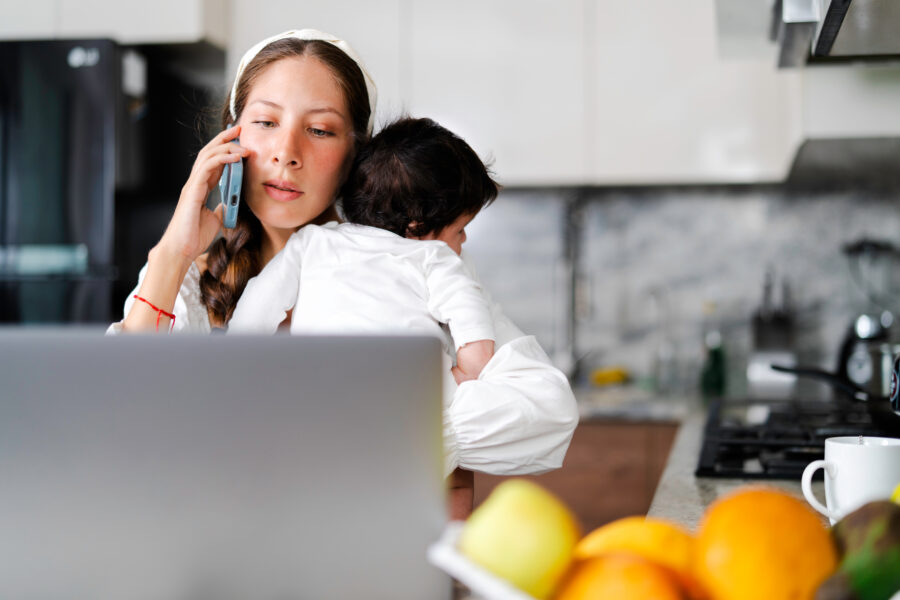 Young mother multitasking in kitchen, working on laptop, holding her baby and talking on the phone