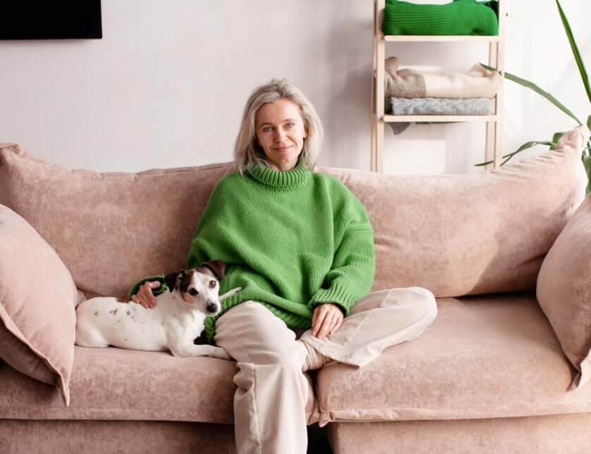 Positive relaxed smiling woman leaning on cozy couch, daydreaming, looking at camera, time alone at home. Lady with dog sitting on sofa at living room