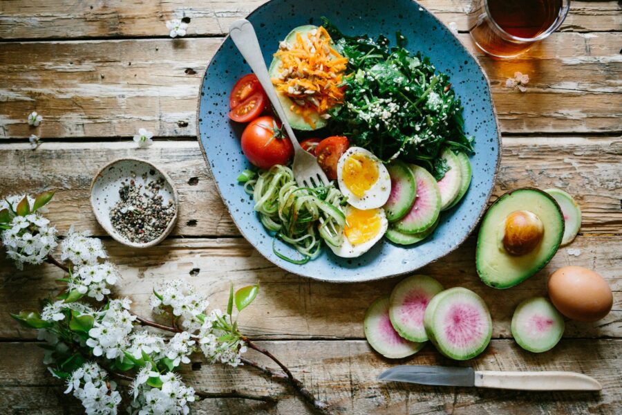 placed on a rustic wooden table is some flowers, a blue bowl filled with colourful foods such as avocardo, tomatos and eggs. Other ingredients are scattered on the table along with a brown drink.