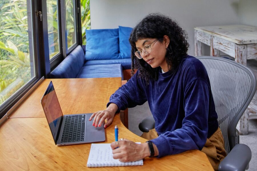 Lady sitting at kitchen table working with a laptop and notepad.