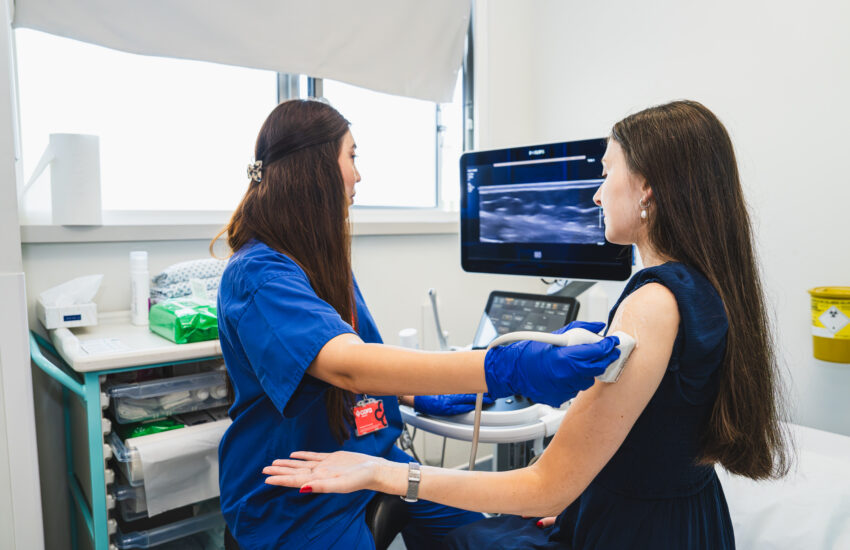 Two ladies in a clinical setting one is in blue scrubs carrying out an ultrasound of the second lady's arm in a black outfit.