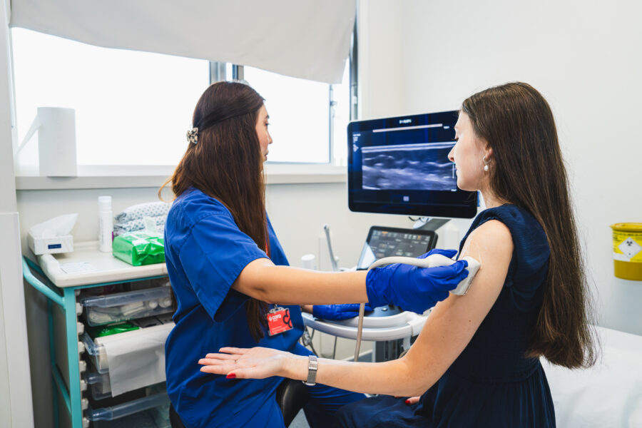Two ladies in a clinical setting one is in blue scrubs carrying out an ultrasound of the second lady's arm in a black outfit.