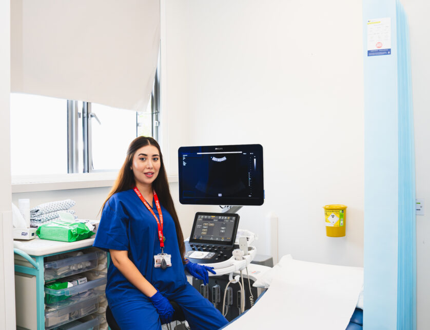 Female clinician sitting in clinical room with blue scrubs on. Behind her is a monitor and Infront a bed and pull around curtain